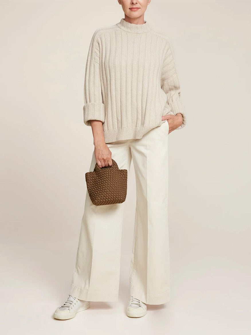 Person in cream-colored sweater and wide-leg white pants holding a Naghedi St. Barths Petit Tote in Solid Mink, featuring a crossbody strap, standing against a neutral background.