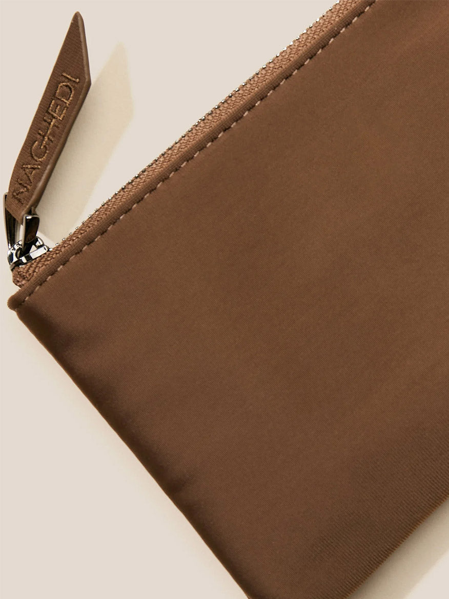 Close-up of a brown zippered pouch with a leather tag displaying the brand name "MACHETE." The handwoven neoprene material adds texture, while the crossbody strap offers versatility. The pouch is set against a neutral background, reminiscent of the chic Naghedi St. Barths Petit Tote in Solid Mink collection.