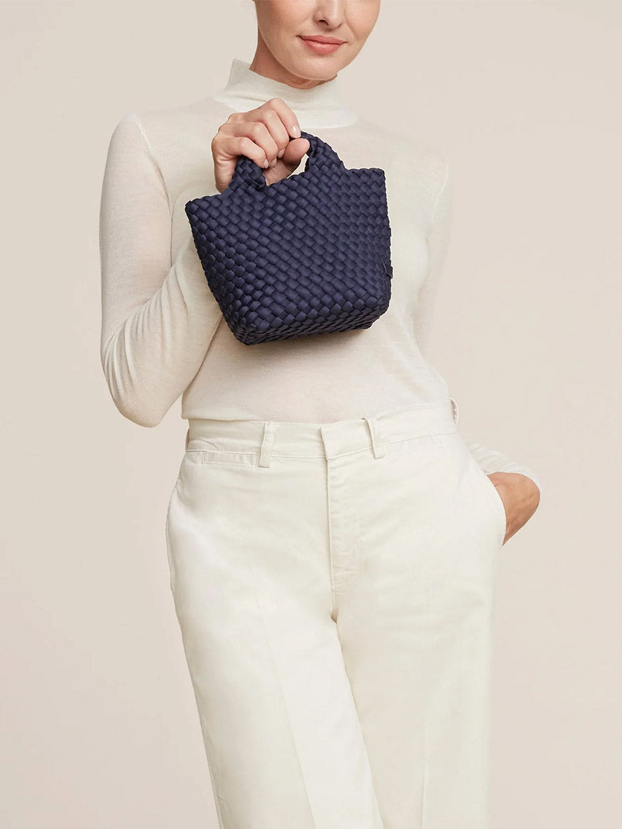 A woman in a beige turtleneck and white pants holding a Naghedi St. Barths Petit Tote in Solid Ink Blue handbag. Only her torso and hand are visible.