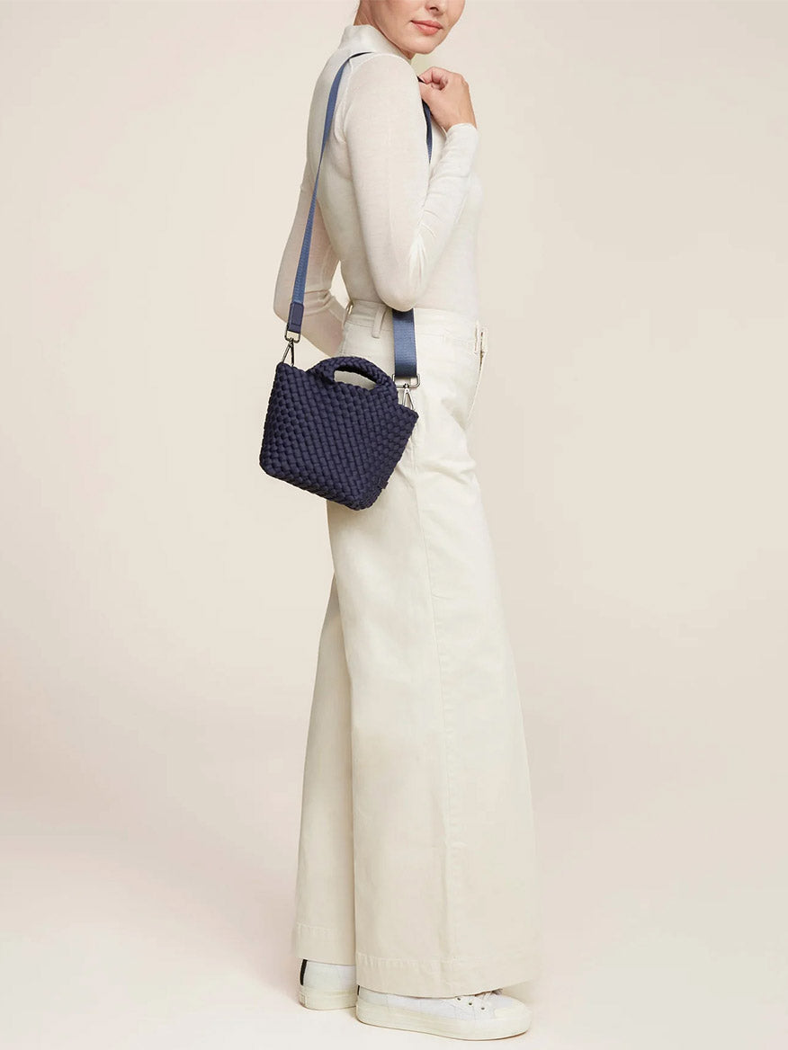 Woman in a white long-sleeve top and wide-leg trousers, carrying a Naghedi St. Barths Petit Tote in Solid Ink Blue with a crossbody strap over her shoulder, standing against a light beige background.