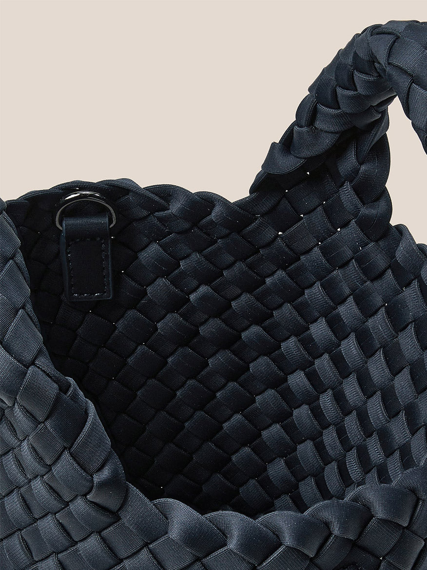 Close-up of a black woven basket with a braided handle, highlighting its interior and featuring a black crossbody strap connected to a metal ring. This handwoven neoprene design mirrors the elegant style of the Naghedi St. Barths Petit Tote in Solid Murano.