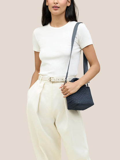 A person dressed in a white t-shirt and cream pants accessorized with a white belt is carrying the sophisticated Naghedi St. Barths Petit Tote in Solid Murano, crafted from handwoven neoprene, on their shoulder.