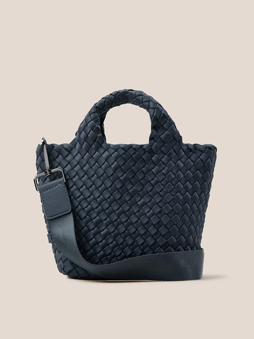 The Naghedi St. Barths Petit Tote in Solid Murano is a small, dark blue bag crafted from handwoven neoprene, featuring short handles and a detachable crossbody strap, showcased against a plain beige background.