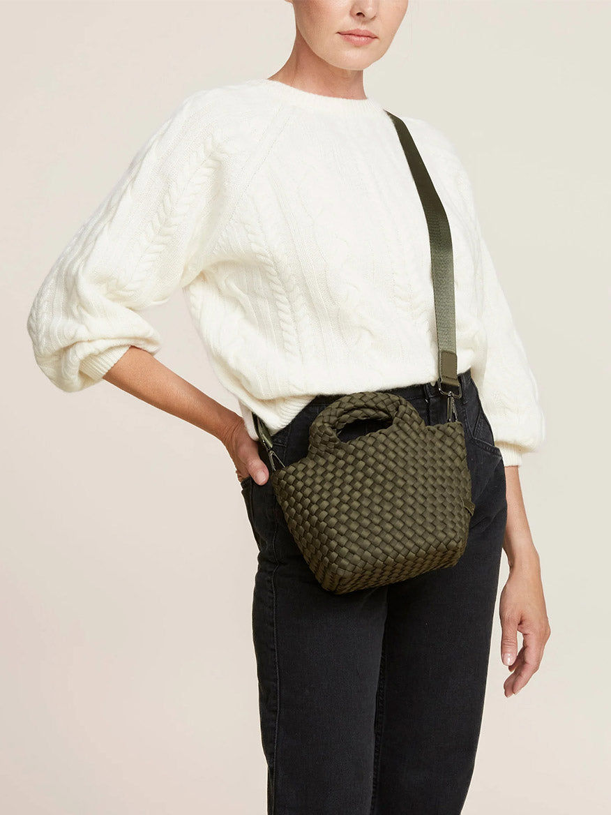 A person wearing a white sweater and black pants carries a Naghedi St. Barths Petit Tote in Solid Olive, featuring a small, handwoven neoprene design with a sleek green crossbody strap.