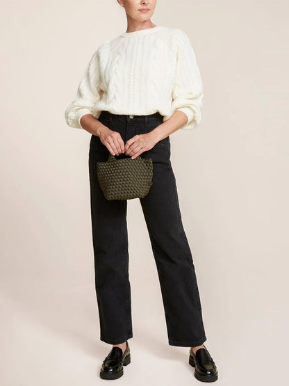 A person wearing a white knitted sweater and black jeans, holding a stylish Naghedi St. Barths Petit Tote in Solid Olive with a crossbody strap, and wearing black loafers.