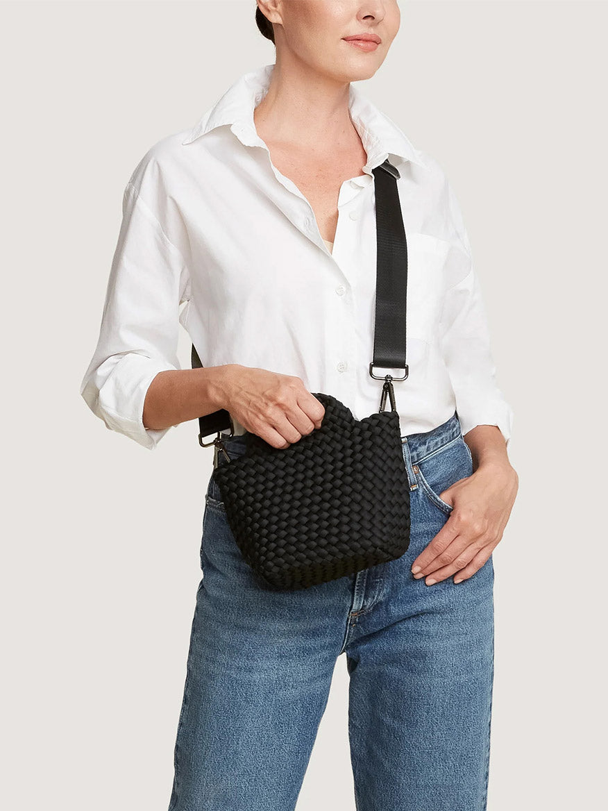 A person in a white button-down shirt and blue jeans carries a black Naghedi St. Barths Petit Tote, a mini handwoven neoprene bag featuring a crossbody strap.