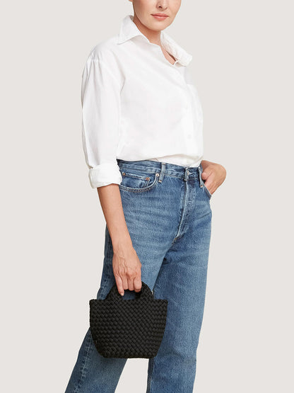 A person wearing a white button-down shirt and blue jeans holds a small black handwoven Naghedi St. Barths Petit Tote in Onyx.