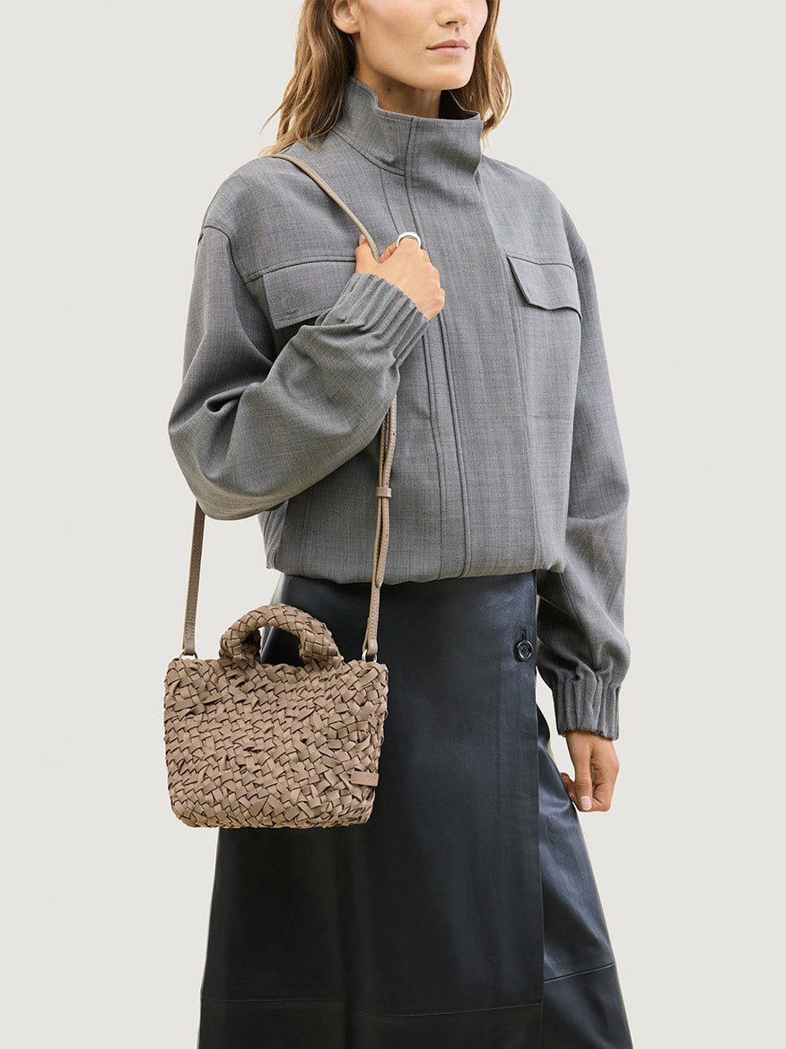 A person wearing a gray jacket and black skirt carries a taupe Naghedi St. Barths Petit Tote Nest Weave in Cashmere, a small, handwoven neoprene bag with a crossbody strap.