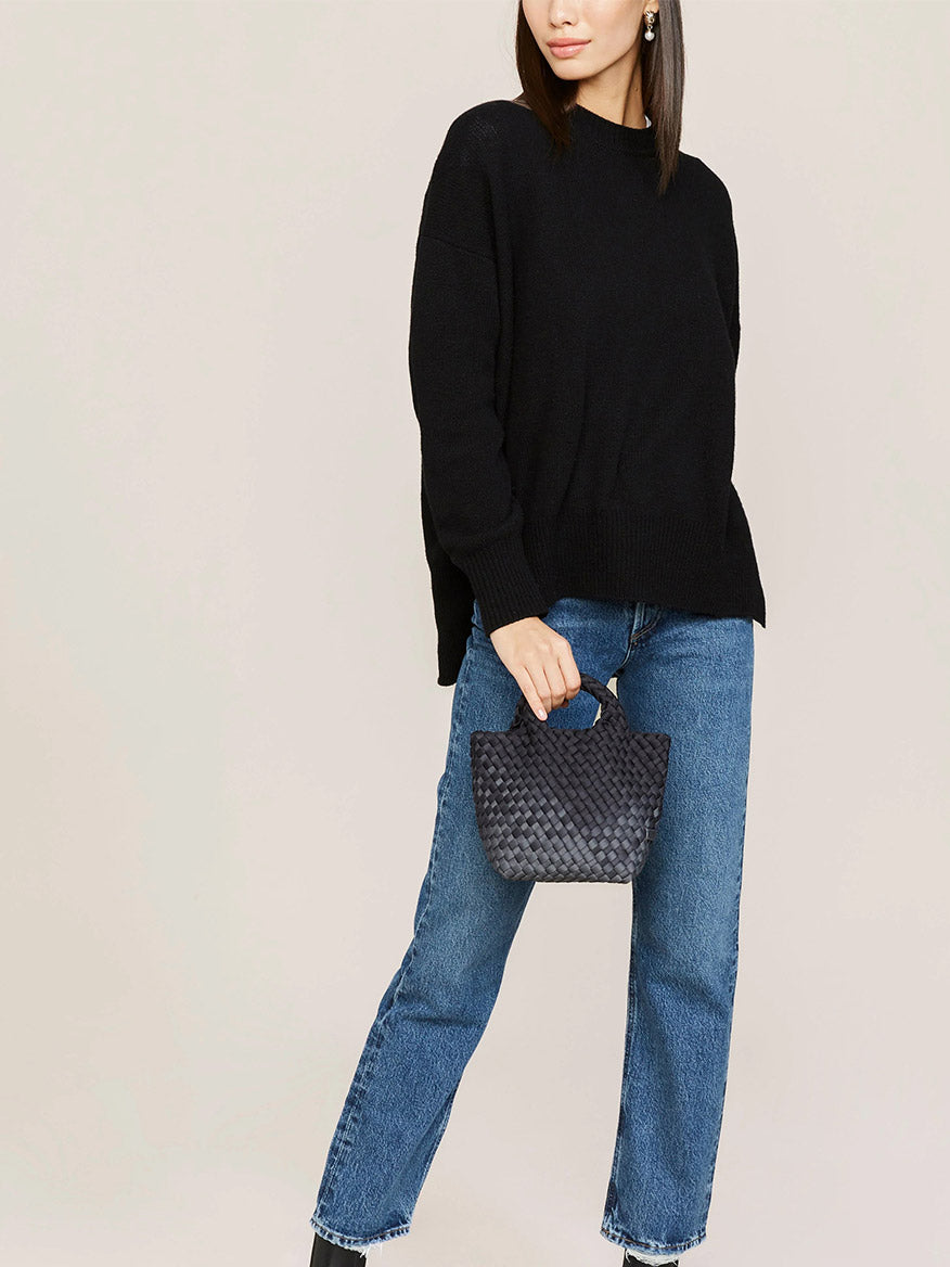 Woman in a black sweater and blue jeans holding a gray handwoven neoprene Naghedi St. Barths Petit Tote in Graphic Ombre Basalt.