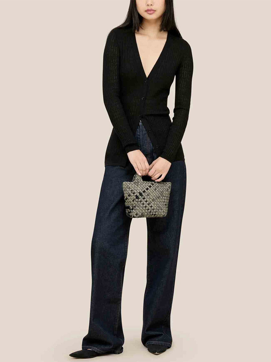 A woman wearing a black cardigan and wide-leg jeans holds a Naghedi St. Barths Petit Tote in Watercolor Neptune, featuring a handwoven neoprene finish, against a plain background.