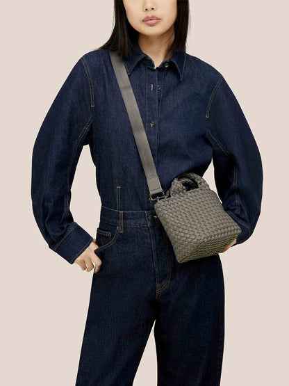 A person in a dark denim outfit holds a Naghedi St. Barths Petit Tote in Solid Laurel with a crossbody strap, standing against a plain background.