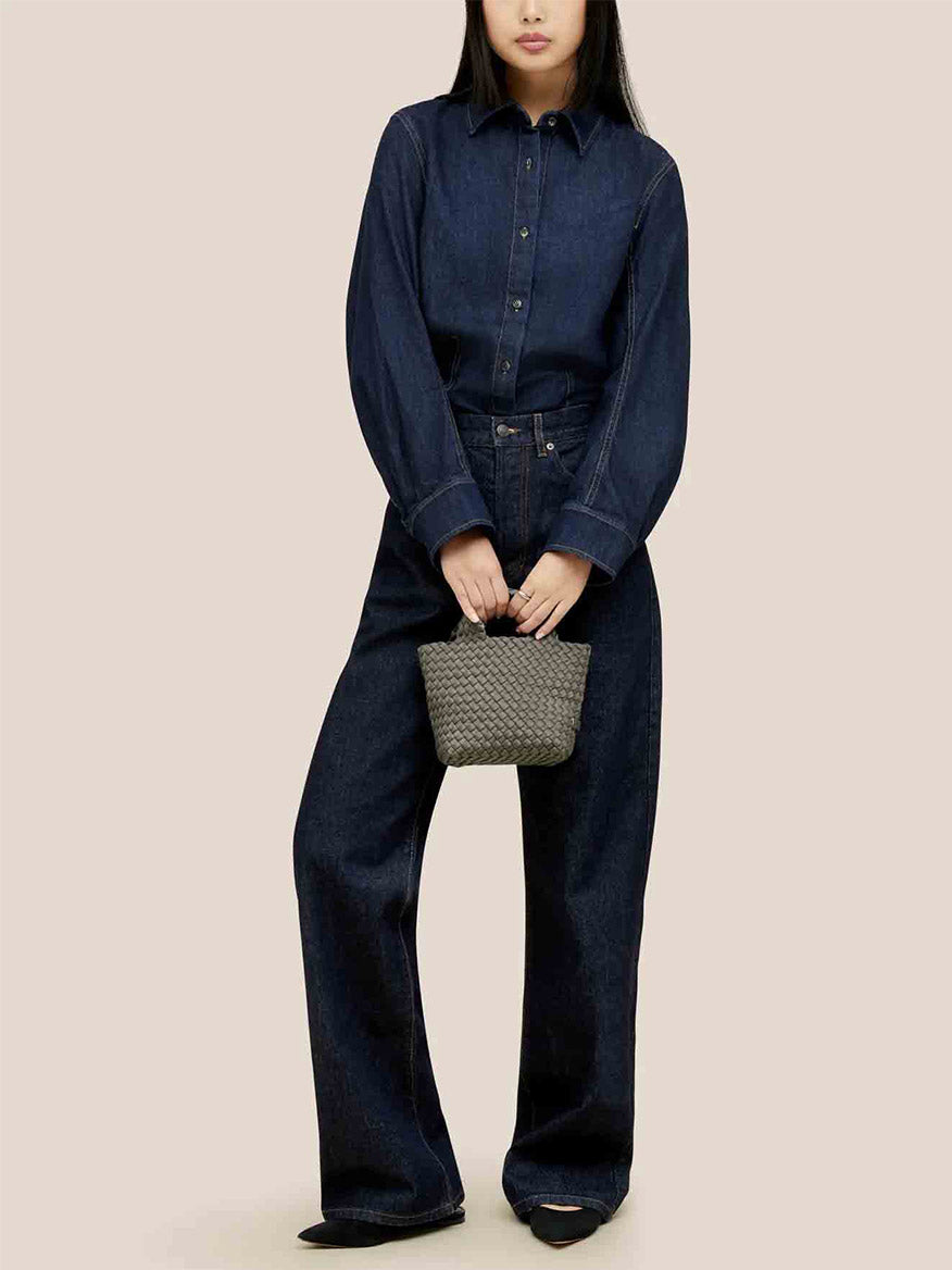 A person dressed in a dark denim outfit is holding a Naghedi St. Barths Petit Tote in Solid Laurel with a crossbody strap, standing against a plain background.