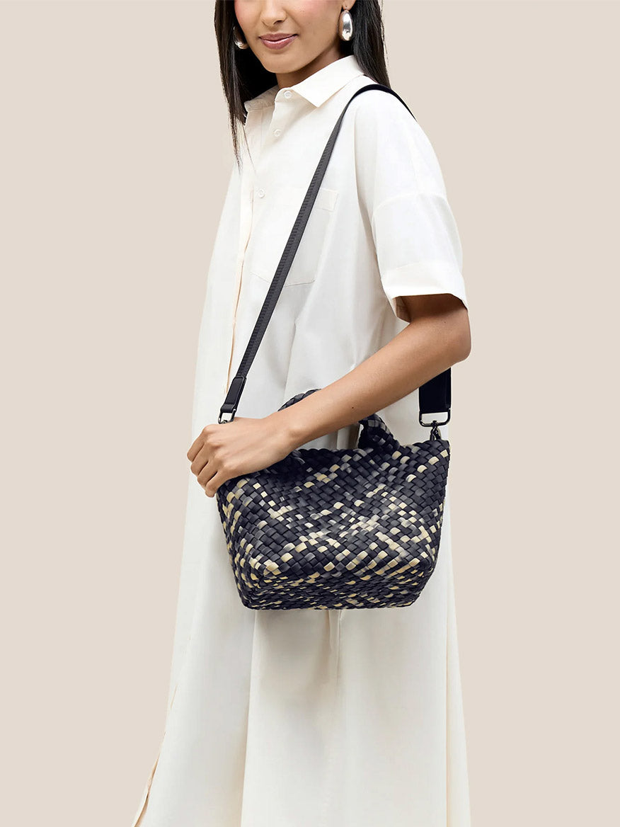 A person in a white dress holds a Naghedi St. Barths Small Tote in Watercolor Nimbus, featuring handwoven neoprene with black and beige accents and a detachable crossbody strap.