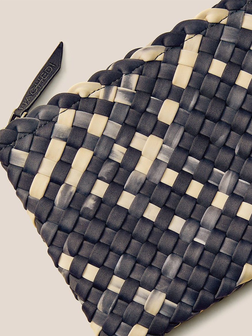 A detailed view of the Naghedi St. Barths Small Tote in Watercolor Nimbus reveals its handwoven neoprene surface with a captivating black and white checker pattern, complete with a charming zipper tag enhancing its appeal.