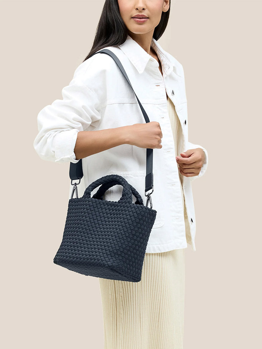 A person dressed in a white jacket and light dress carries the Naghedi St. Barths Small Tote in Solid Murano, a handwoven black neoprene handbag featuring a crossbody strap.