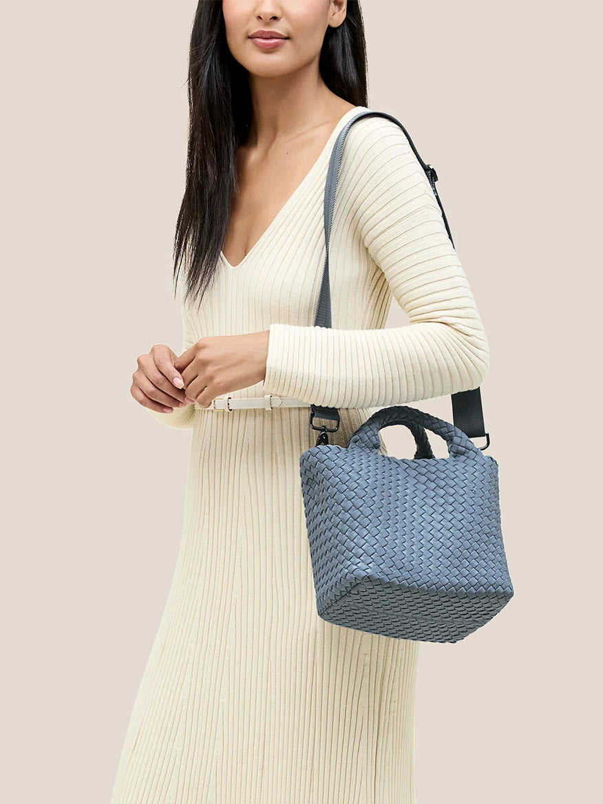 A woman in a long cream ribbed dress holds a Naghedi St. Barths Small Tote in Solid Nova with a crossbody strap, its handwoven neoprene design accentuating her style against a plain background.