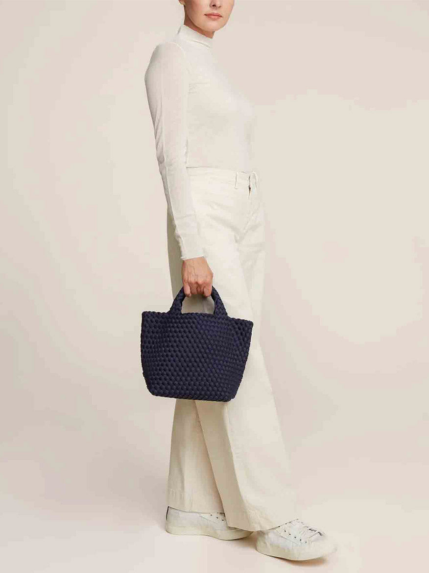 A person in a white outfit is holding the Naghedi St. Barths Small Tote in Solid Ink Blue, featuring a handwoven design and crossbody strap, while standing against a plain background.