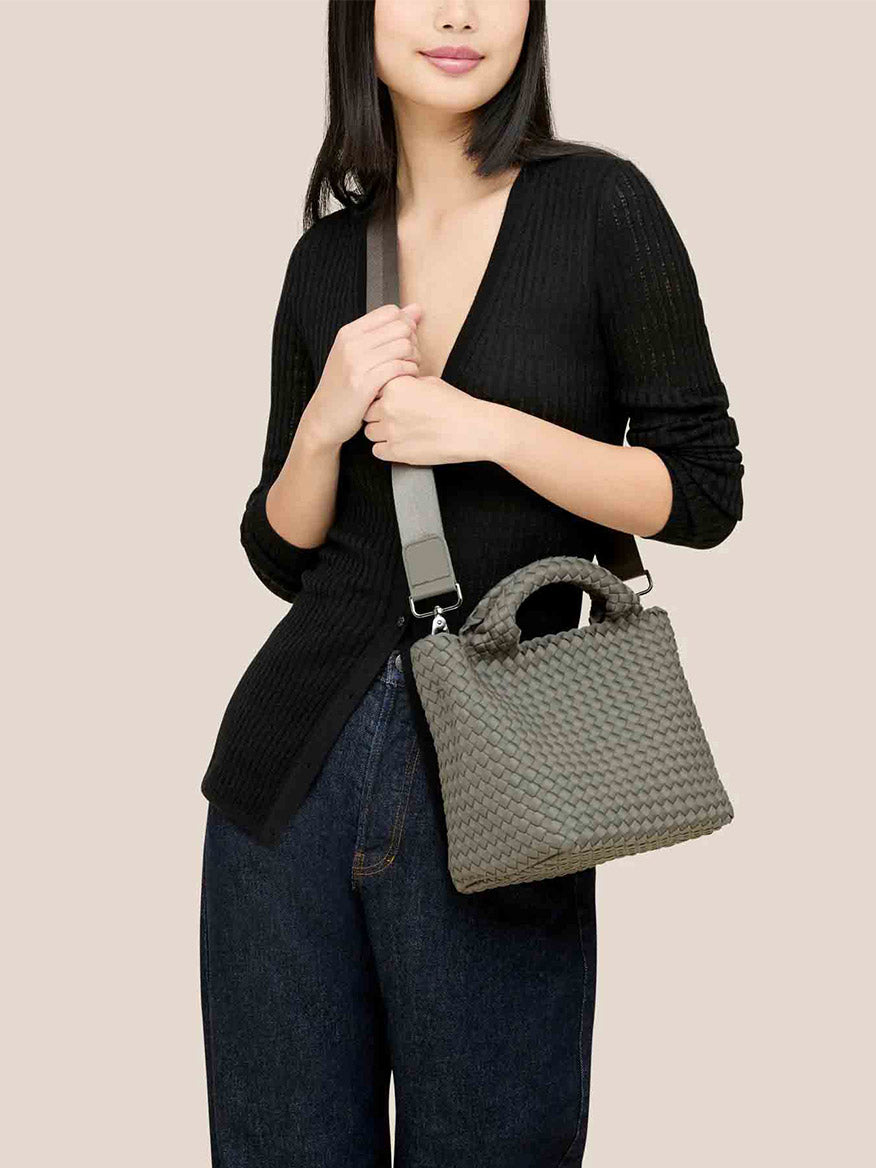 A person in a black sweater and jeans holds the Naghedi St. Barths Small Tote in Solid Laurel, showcasing its textured gray design with an adjustable crossbody strap, against a plain background.