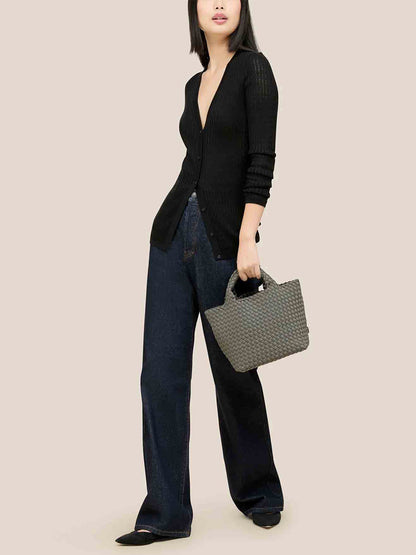 Wearing a black sweater and dark wide-leg jeans, a person holds the Naghedi St. Barths Small Tote in Solid Laurel with an adjustable crossbody strap, styled against a neutral background.