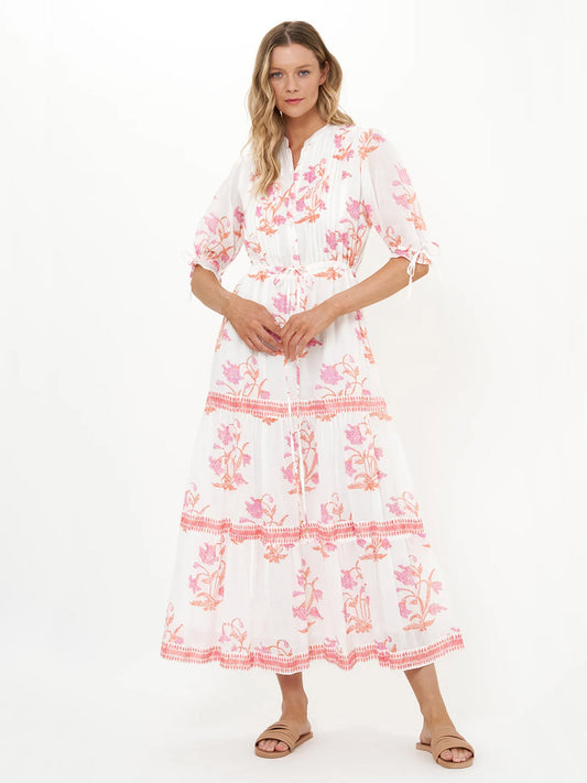 A woman wears the Oliphant Cinched Pintuck Maxi Dress in Ostuni Coral, featuring hand-block prints, white cotton voile fabric, pink floral patterns, and three-quarter sleeves, paired with brown flat sandals.