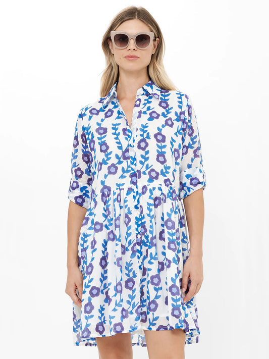 A woman models the Oliphant Long Sleeve Shirt Dress Mini in Toscana Blue, featuring blue and purple floral patterns, sunglasses, and a chic white backdrop—blending effortless style with comfort.