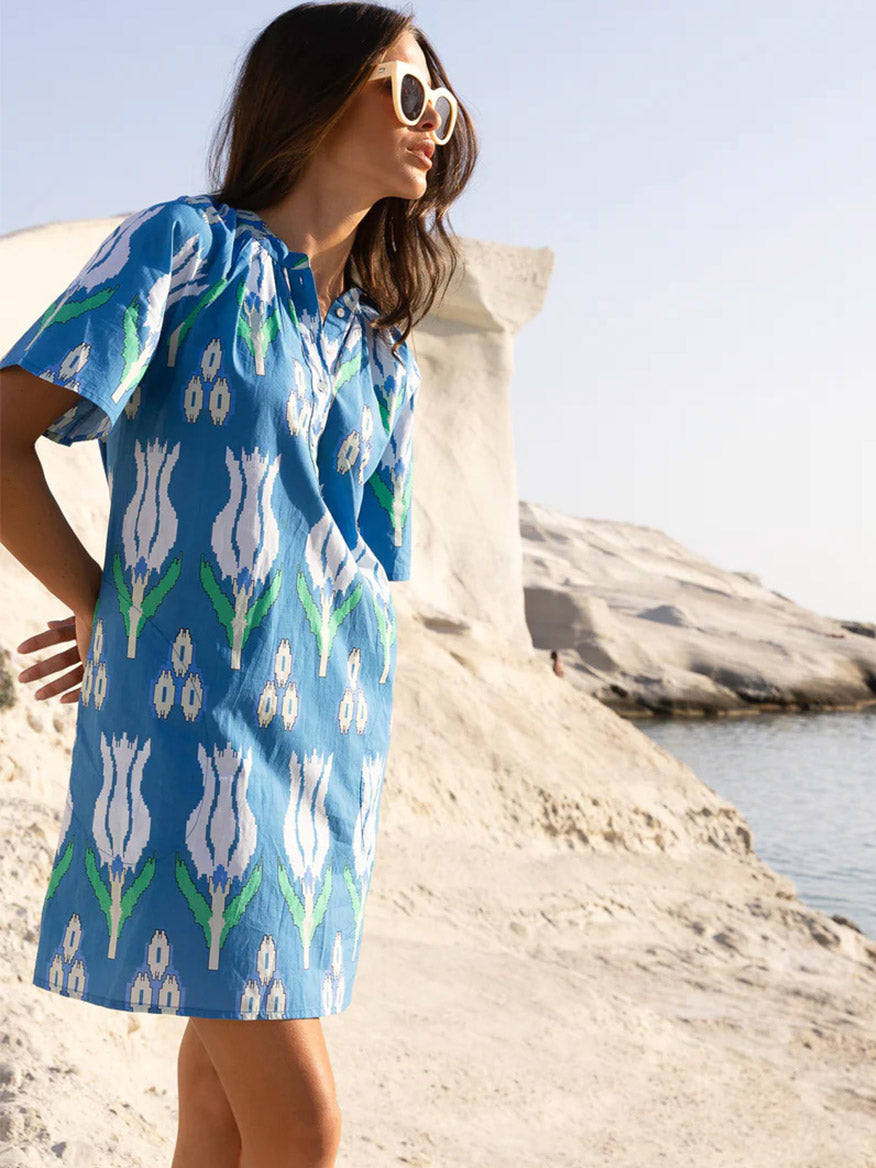 Woman posing on a rocky beach while wearing an Oliphant Pocket Dress in Sumba Blue Tulip featuring pockets and white sunglasses.