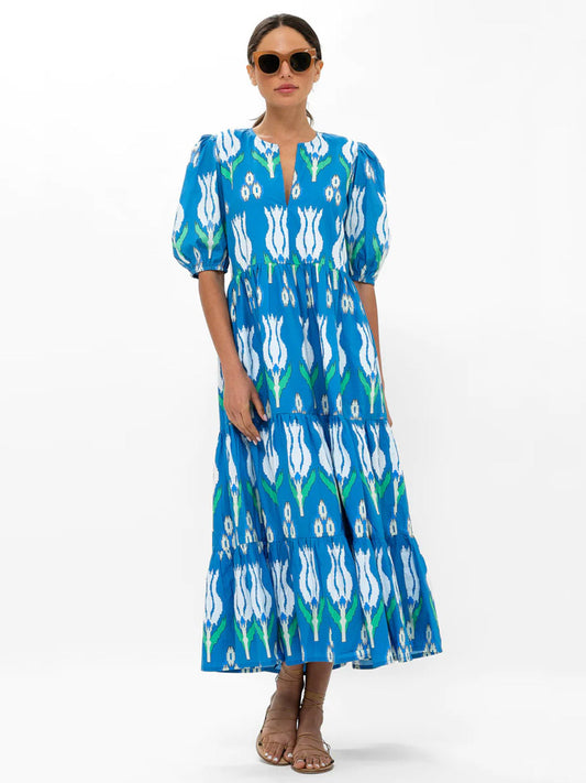 A woman modeling an Oliphant Puff Sleeve Maxi Dress in Sumba Blue Tulip, paired with sunglasses and brown sandals.