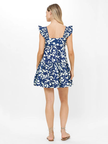 A woman stands with her back to the camera, wearing the Oliphant Smocked Square Neck Mini Dress in Acadia Navy paired with flat sandals, set against a plain white background.