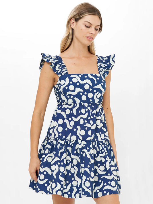 A woman wears the Oliphant Smocked Square Neck Mini Dress in Acadia Navy, featuring blue and white abstract print, tiered A-line silhouette, ruffle details; standing against a plain white background.