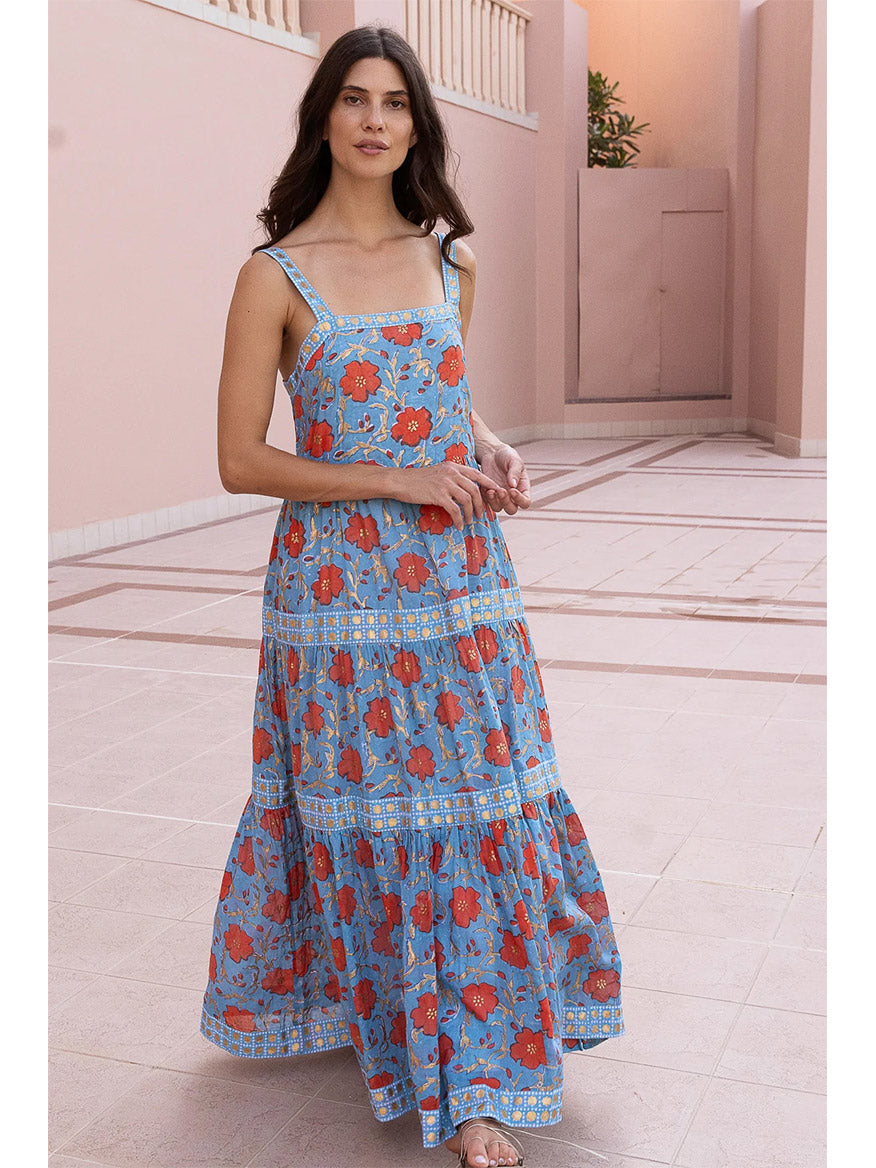 A woman stands in a pink corridor wearing an Oliphant Strappy Maxi Dress in Poppy Red with gold paint detailing, looking directly at the camera.