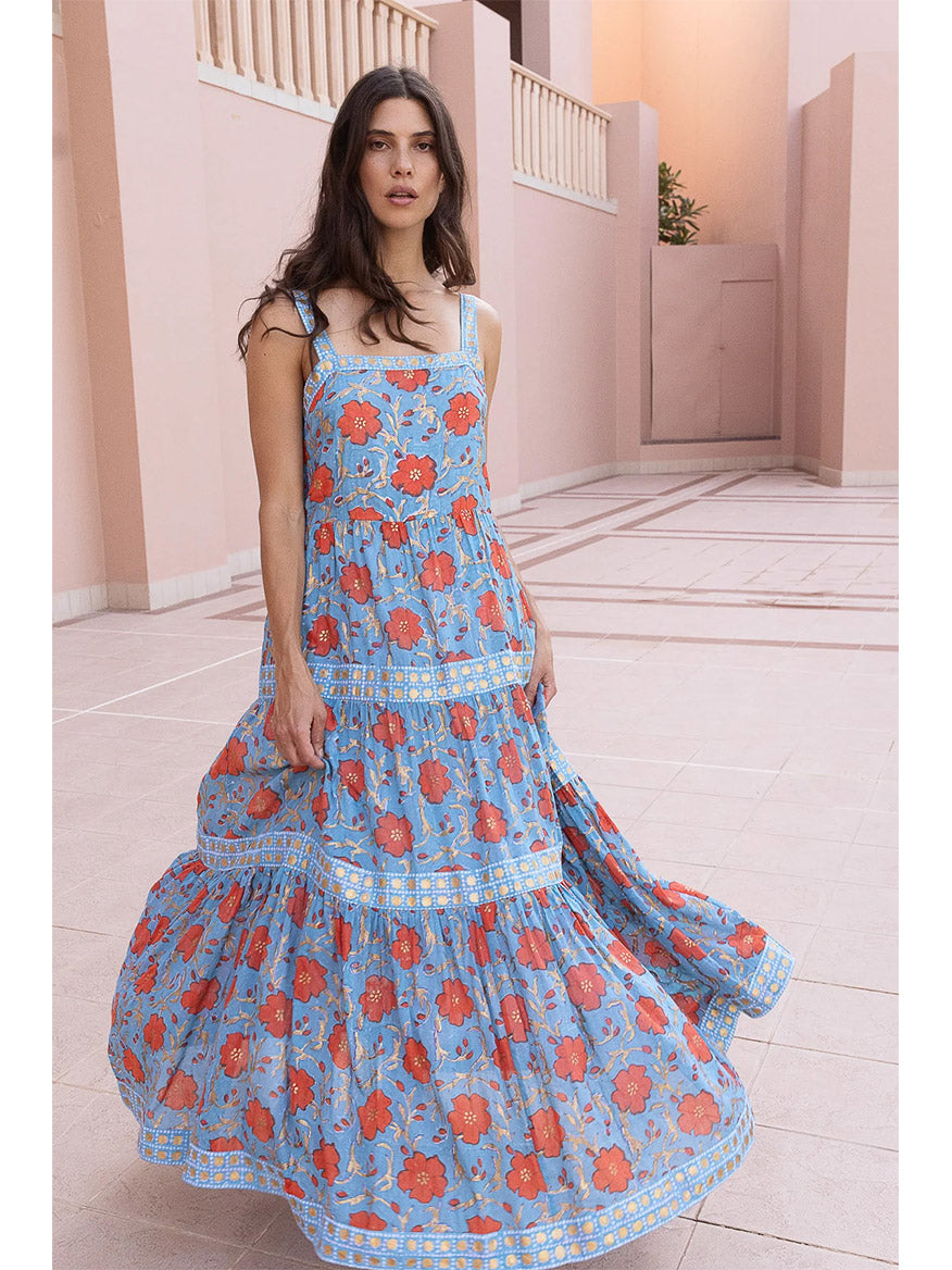 A woman in an Oliphant Strappy Maxi Dress in Poppy Red stands in a pink corridor with arches.