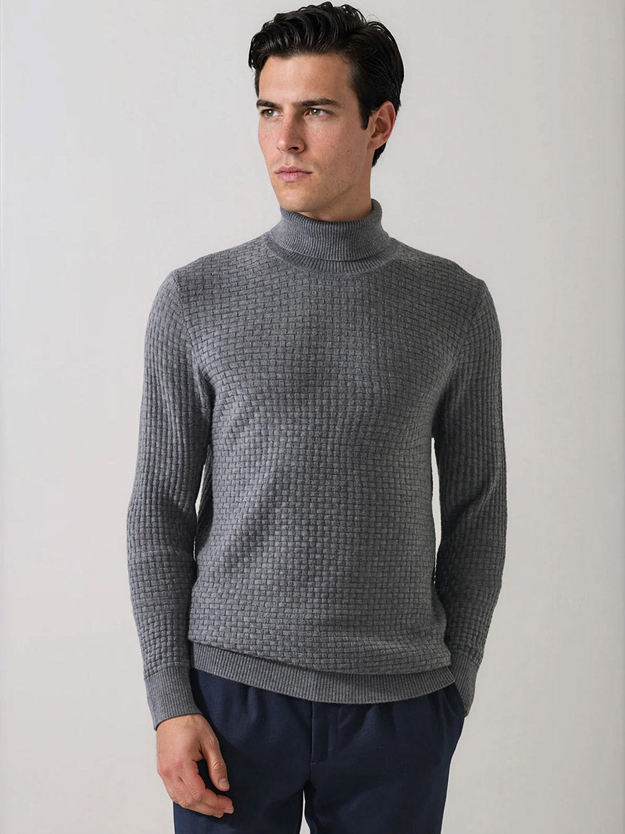 A man wearing the Patrick Assaraf Extra-Fine Merino Wool Basketweave Turtleneck in Castlerock and dark pants stands against a plain light background, looking to the side. The distinctive basketweave knit of this merino wool sweater adds visual interest and texture to his outfit.