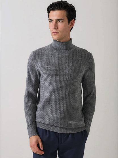 A man wearing the Patrick Assaraf Extra-Fine Merino Wool Basketweave Turtleneck in Castlerock and dark pants stands against a plain light background, looking to the side. The distinctive basketweave knit of this merino wool sweater adds visual interest and texture to his outfit.