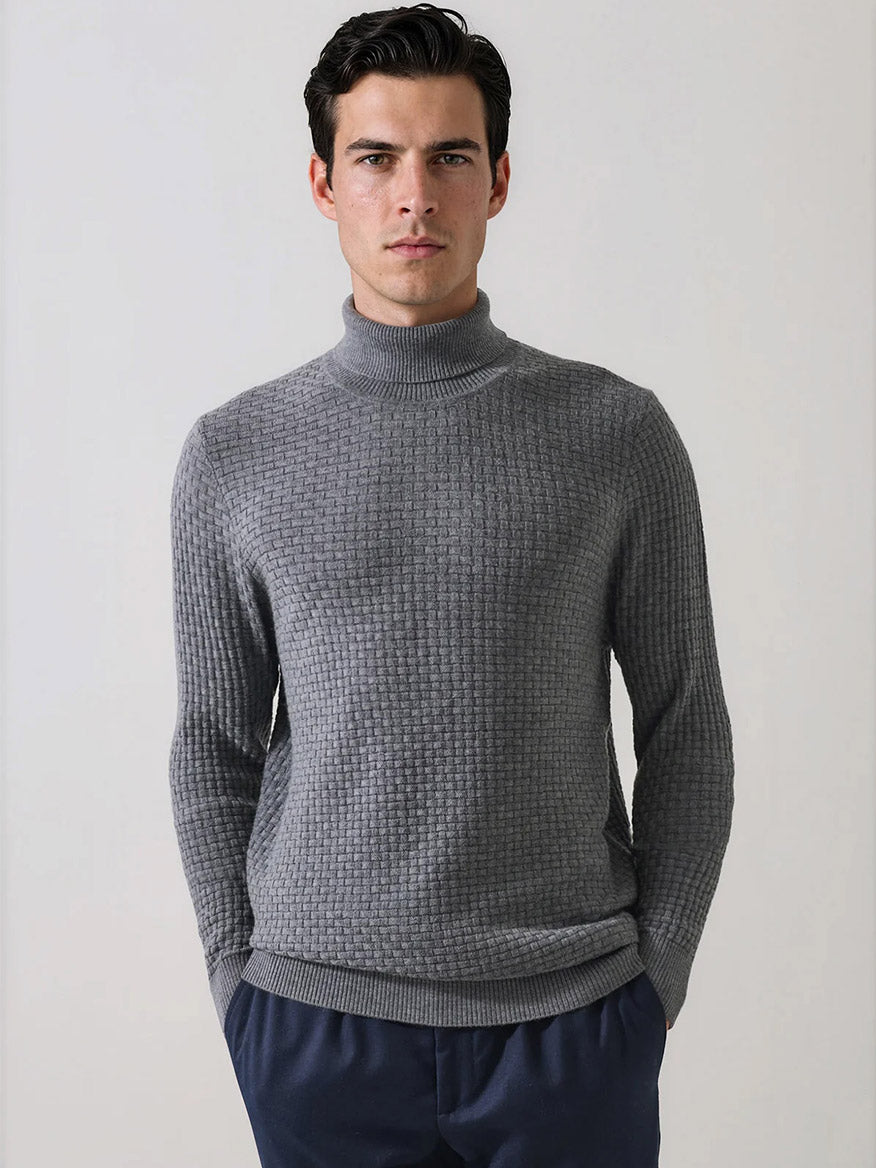 A man stands against a plain background wearing the Patrick Assaraf Extra-Fine Merino Wool Basketweave Turtleneck in Castlerock, its distinct basketweave knit adding refined texture to his look. He pairs this sophisticated layering sweater with navy blue pants, gazing directly at the camera with his hands in his pockets.