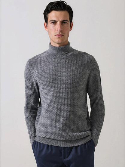 A man stands against a plain background wearing the Patrick Assaraf Extra-Fine Merino Wool Basketweave Turtleneck in Castlerock, its distinct basketweave knit adding refined texture to his look. He pairs this sophisticated layering sweater with navy blue pants, gazing directly at the camera with his hands in his pockets.