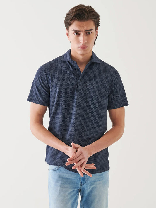 A young man with brown hair stands against a white background in light blue jeans and the Patrick Assaraf Pima Cotton Stretch Printed Polo in Midnight, his hands clasped in front.