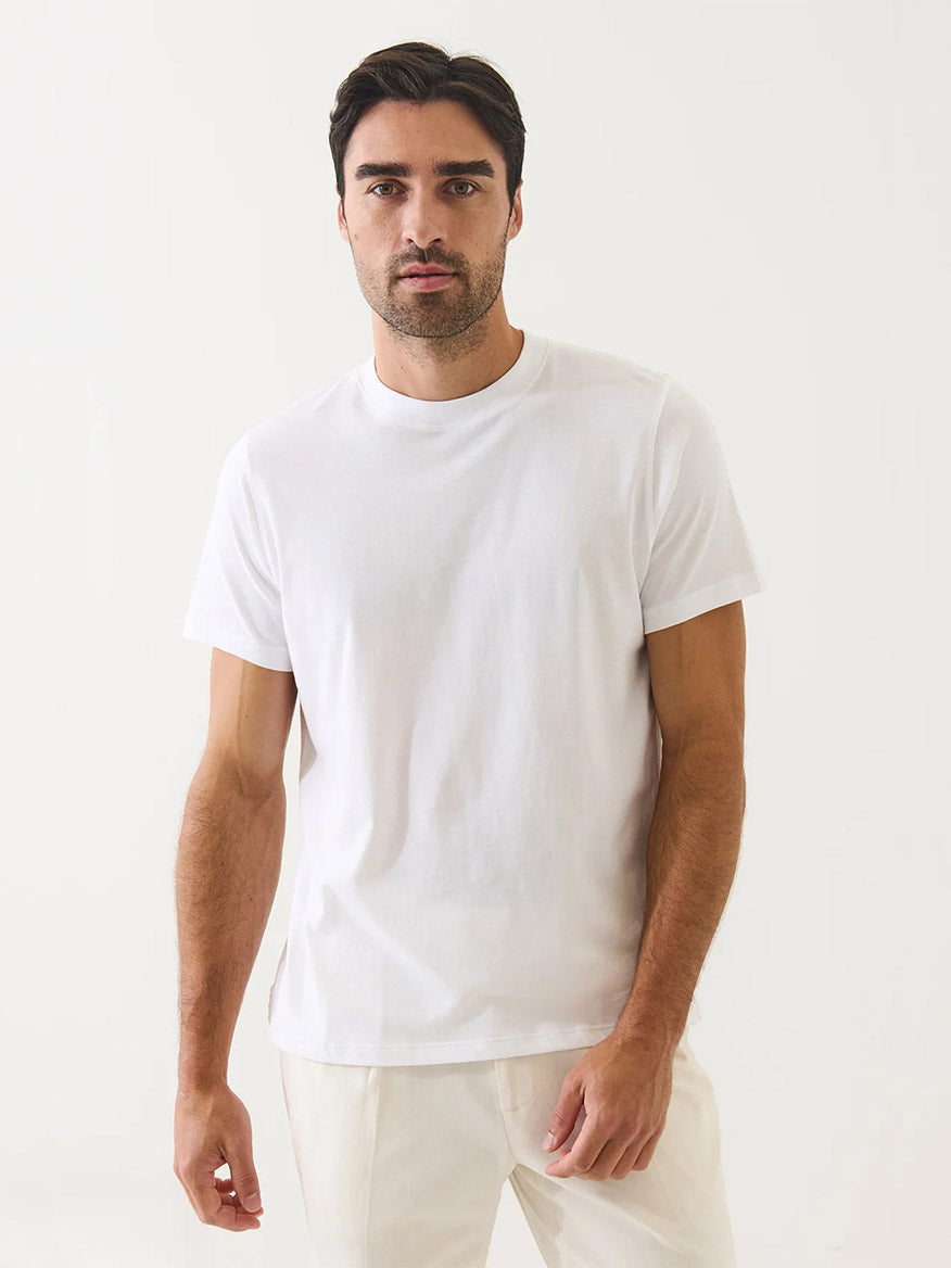 A man with dark hair and a beard wears the Patrick Assaraf Organic Pima Cotton High Neck T-Shirt in white and matching pants, standing against a light background—a stylish nod to sustainable fashion.