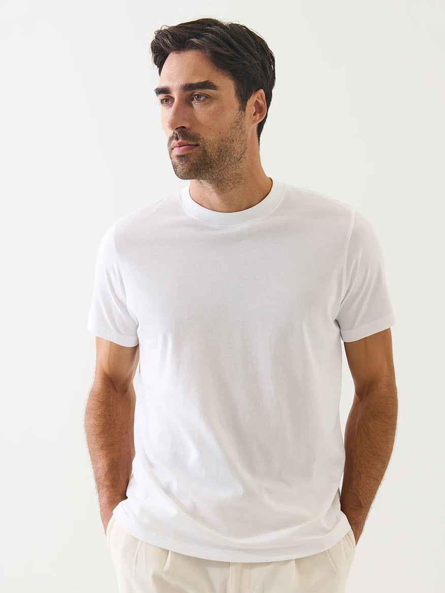 A man with dark hair and light facial hair wears the Patrick Assaraf Organic Pima Cotton High Neck T-Shirt in White and light pants, standing against a plain white background with his hands in his pockets.