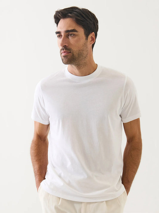 A man with dark hair and light facial hair wears the Patrick Assaraf Organic Pima Cotton High Neck T-Shirt in White and light pants, standing against a plain white background with his hands in his pockets.