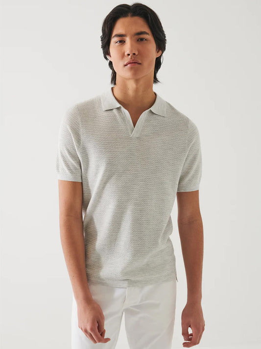 A young man stands wearing a Patrick Assaraf Cotton Cupro Open Polo in Drizzle and white pants against a plain white background.