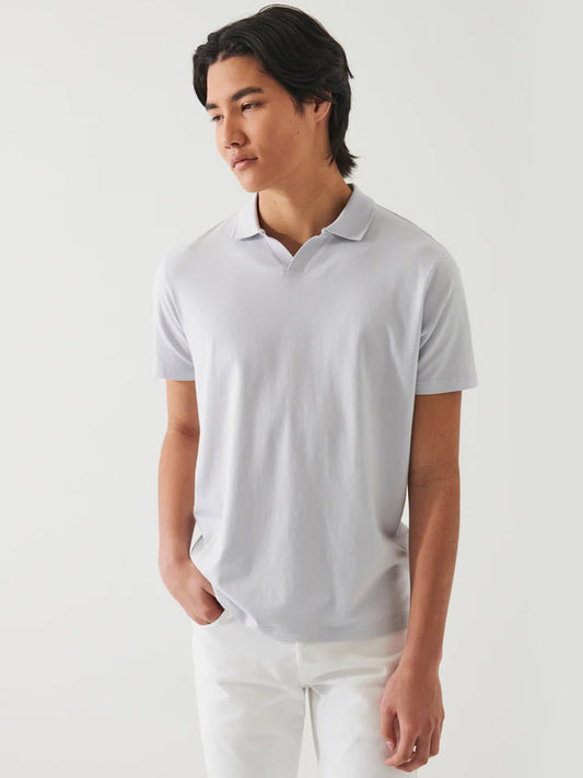 A young man wearing a Patrick Assaraf Pima Cotton Stretch Open Polo in Glacier and white pants, standing against a plain background.