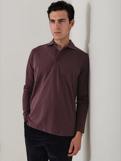 A man stands indoors leaning against a white wall with his hands in his pockets, wearing black pants and the Patrick Assaraf Pima Cotton Stretch Long Sleeve Polo in Prune, crafted from luxurious Peruvian Pima cotton, and gazes to the side.