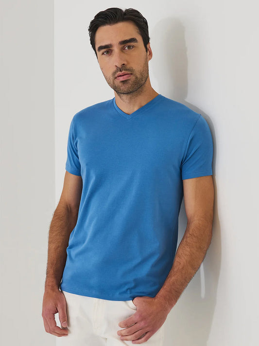 A man with dark hair and a short beard wears the Patrick Assaraf Pima Cotton Stretch V-Neck T-Shirt in Harbour with white pants, standing against a white wall, his left hand in his pocket.