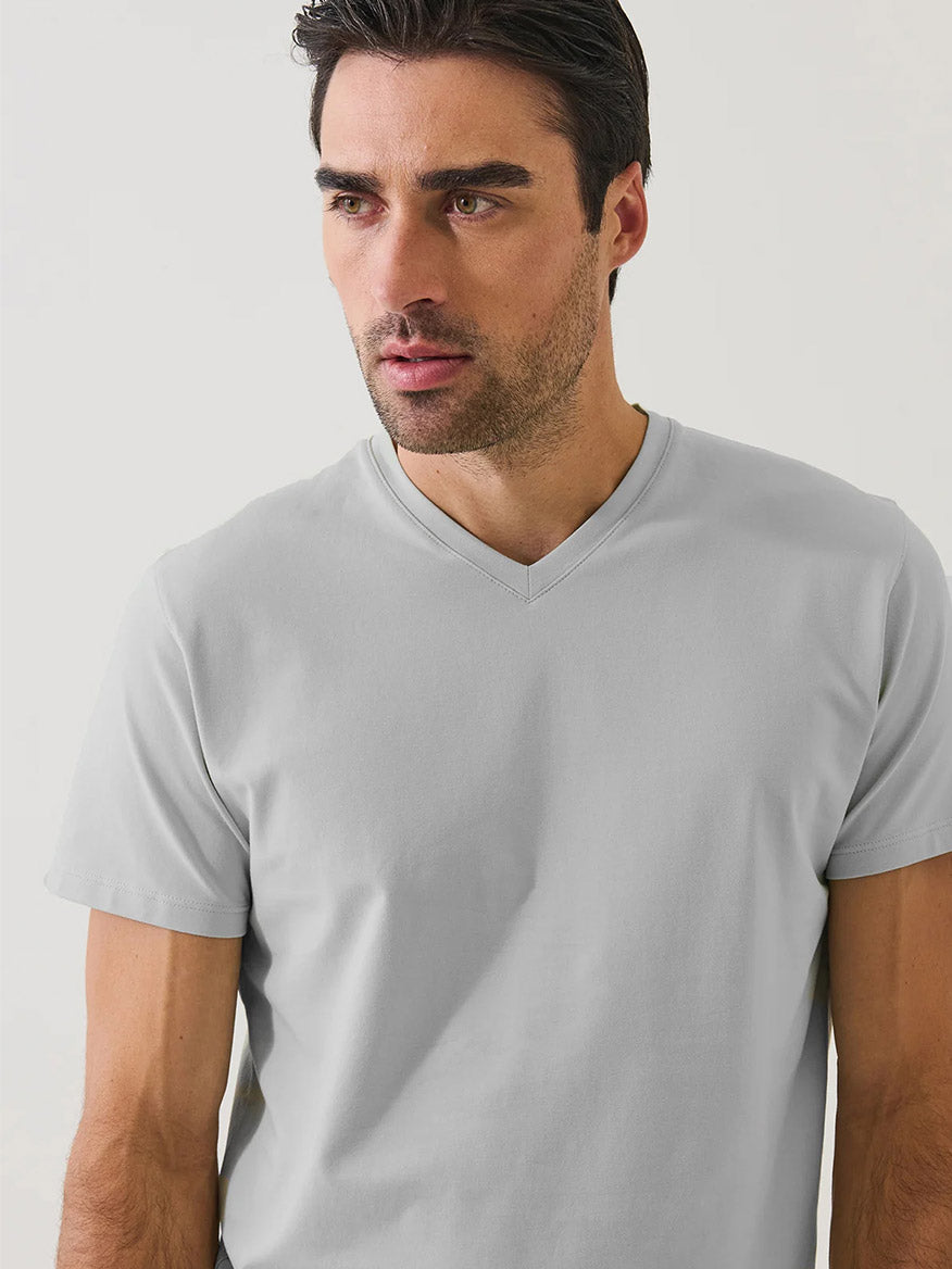 A man with dark hair and stubble wears the Patrick Assaraf Pima Cotton Stretch V-Neck T-Shirt in Mellow Grey, standing against a plain white background and looking slightly to the side.