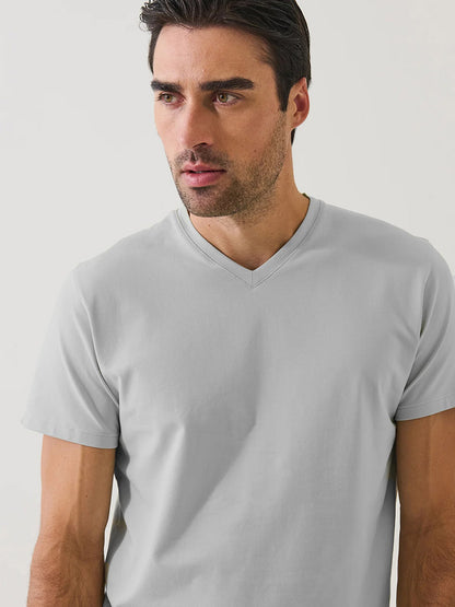 A man with dark hair and stubble wears the Patrick Assaraf Pima Cotton Stretch V-Neck T-Shirt in Mellow Grey, standing against a plain white background and looking slightly to the side.