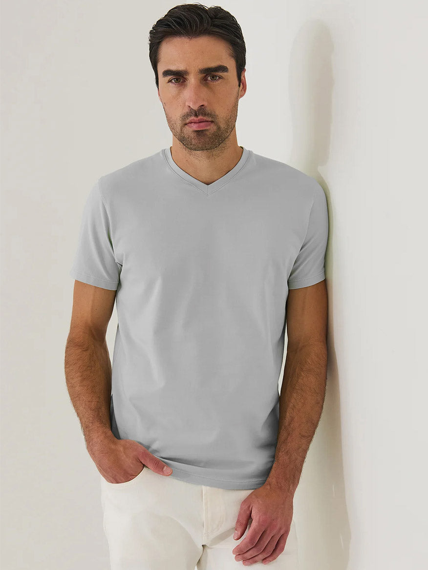 A man with short dark hair and light stubble wears a Patrick Assaraf Pima Cotton Stretch V-Neck T-Shirt in Mellow Grey and white pants, standing against an off-white wall with one hand in his pocket.