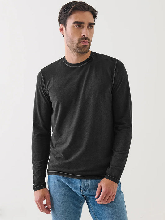 A man with dark hair and facial hair wears the Patrick Assaraf Pima Cotton Stretch Vintage Wash Long Sleeve T-Shirt in Black, paired with blue jeans, standing against a plain white background.