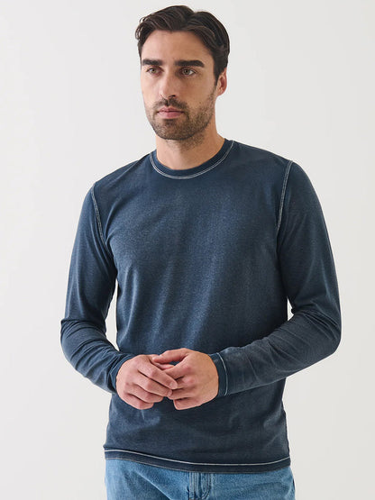A man with short dark hair and a beard wears the Patrick Assaraf Pima Cotton Stretch Vintage Wash Long Sleeve T-Shirt in Midnight, paired with blue jeans, standing against a plain white background.