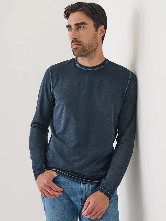 A man wearing the Patrick Assaraf Pima Cotton Stretch Vintage Wash Long Sleeve T-Shirt in Midnight and jeans stands against a white wall with one hand in his pocket, looking to the side.