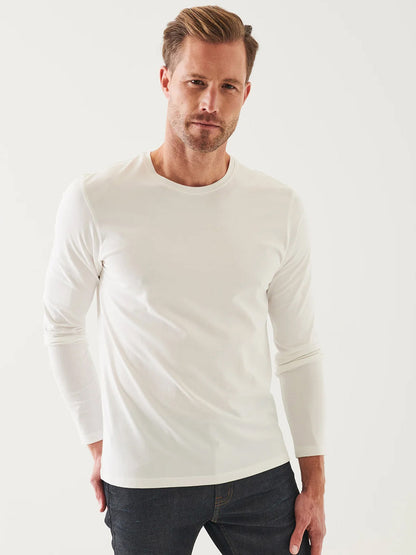 A man wearing a Patrick Assaraf Pima Cotton Stretch Long Sleeve T-Shirt in White and dark jeans stands against a plain light background, looking directly at the camera.